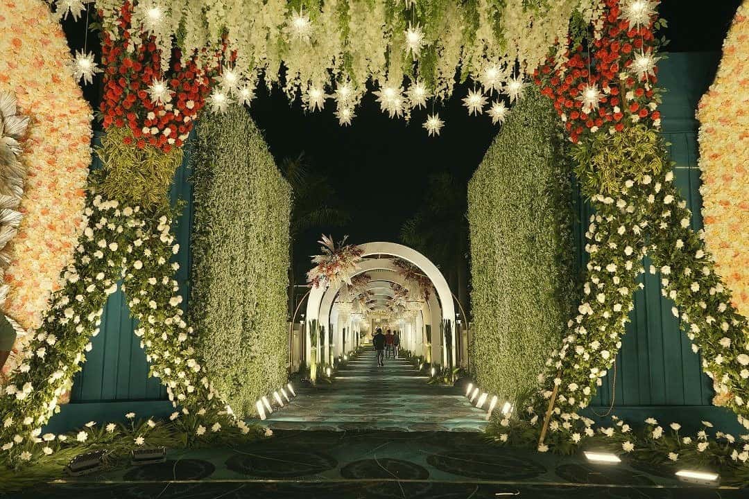 A lit walkway of white flowers leading through a row of arches — the way into a celebration at The Royal Residency