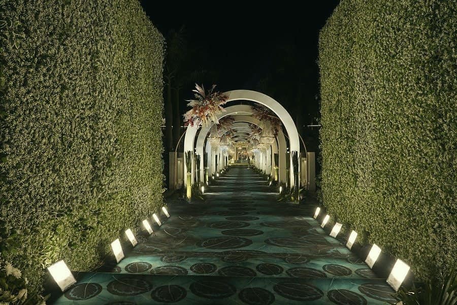 A lit walkway between tall green hedges leading to white arches